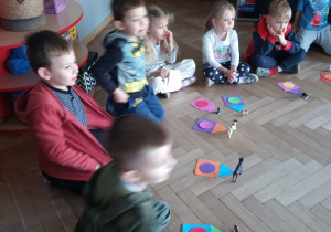 Dzieci siedzą a przed nimi ułożone w odpowiedni sposób figury geometryczne i figurki psów.