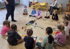 Zdjęcie przedstawia dzieci siedzące w półkolu. Po środku leżą zabawki dla psa.