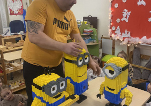 Zdjęcie przedstawia mężczyznę pokazującego dzieciom Minionki z Lego.