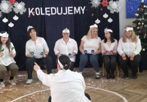 sześć kobiet siedzi przed napisem kolędujemy, w rękach trzymają dzwonki diatoniczne, przed nimi, na podłodze siedzi kobieta