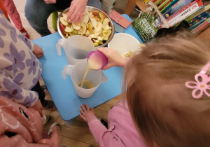 Zdjęcie przedstawia dziewczynkę, wlewającą do dzbanka sok jabłkowy.