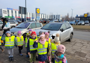 Zdjęcie przedstawia grupkę dzieci pozującą do zdjęcia. Woddali widnieje sklep Lidl.