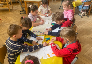 Zdjęcie przedstawia dzieci siedzące przy stoliku, wykonują prace plastyczną.