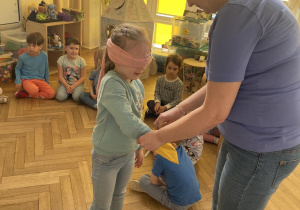 Zdjęcie przedstawia kobietę trzymającą woreczek, do którego wkłada rękę dziewczynka z zasłoniętymi oczami.