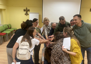 Zdjęcie przedstawia grupę osób stojących w kole. Rodzice mają wyciągniętą rękę przed siebie . Układają ręce jedna na drugą.