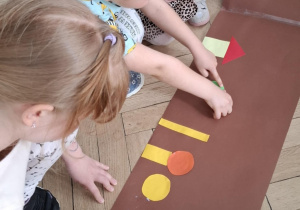 dzieci przyklejają figury geometryczne na papier, tworzą wspólną pracę