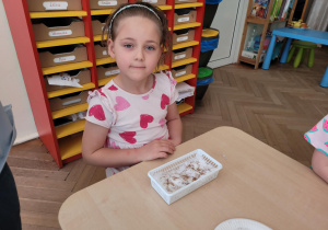 Zdjęcie przedstawia dziewczynkę pozującą do zdjęcia. Dziewczynka ma przed sobą pojemnik z posianymi ziołami.