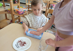 Zdjęcie przedstawia dzieci siedące przy stoliku. Dzieci wsypują nasiona do pojemnika.