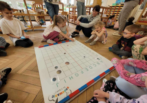 Zdjęcie przedstawia dzieci siedzące wokół maty do kodowania. Dziewczynka stawia na macie robota.