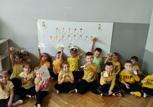 Na zdjęciu widzimy dzieci, które w rękach trzymają wykonane z buletek pszczoły. Za nimi na tablicy widznieje napis "Dzień Pszczoły".