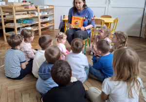 kobieta trzyma w rękach książkę, przed nią siedzą dzieci