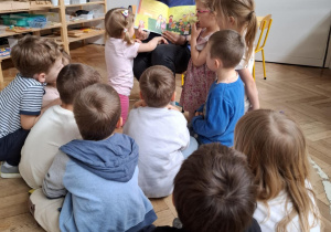 kobieta trzyma w rękach książkę, pokazuje ilustrację w książce,przed nią siedzą dzieci