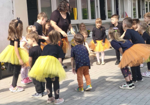 Zdjęcie przedstawia dzieci i nauczycielkę tańczących na tarasie. Wszyscy mają na sobie czarne stroje a dziewczynki dodatkowo żółte lub pomarańczowe sukienki.