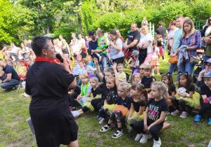 Zdjęcie przedstawia nauczycielkę przemawiającą przez mikrofon do publiczności zgromadzonej w ogrodzie.