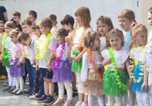 Zdjęcie przedstawia dzieci ubrane w białe koszulki, dziewczynki w kolorowe spódniczki. Wszyscy mają hawajskie naszyjniki.