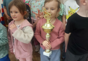 Na zdjęciu dziewczynka, która trzyma puchar, a w tle są dzieci