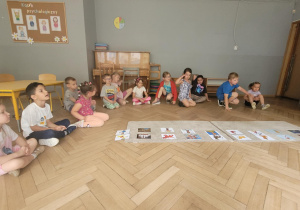 Zdjęcie przedstawia dzieci siedzące na podłodze w półkolu. Przeds dzicmi leżą dywaniki na których zanjdują się zdjęcia psów.