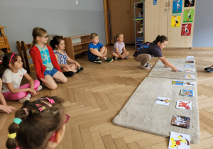 Zdjęcie przedstawia dzieci siedzące na podłodze w półkolu. Przeds dzicmi leżą dywaniki na których zanjdują się zdjęcia psów.