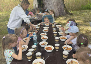 Zdjęcie przedstawia dzieci jedzące spagetti przy stole obok ogniska.