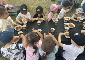 Zdjęcie przedstawia dzieci jedzące spagetti przy stole obok ogniska.