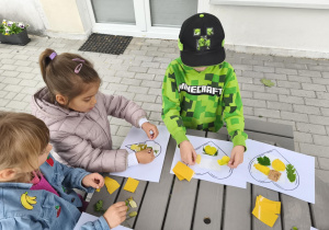 Na zdjęciu widzimy dzieci, które przyklejają materiał naturalny na szablony.