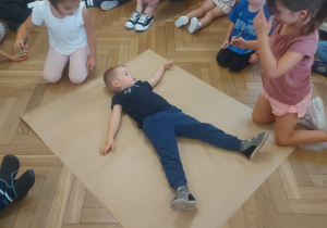 Dzieci zebrane w kole. Dwoje dzieci odrysowuje młodszego kolegę. Tworzymy zasady