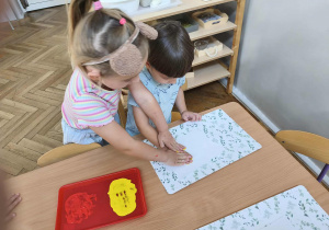 Na zdjęciu widzimy chłopca, który odciska dloń zamoczoną w farbie na kartkę papieru. Jego koleżanka troszkę mu pomaga.
