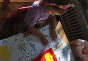 Na zdjęciu widzimy dziewczynkę, która odciska dłoń pomalowaną farbą na kartkę papieru.