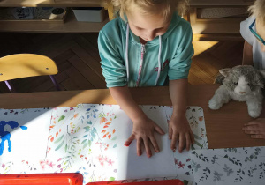 Na zdjęciu widzimy dziewczynkę, która odciska dłoń zamoczoną w farbie na kartkę papieru.