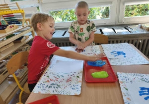Na zdjęciu widzimy dwójkę chłopców. Jeden z nich dciska dłoń umoczoną w farbie na kartkę, a drugi chłopiec macza dłoń w farbie koloru niebieskiego.