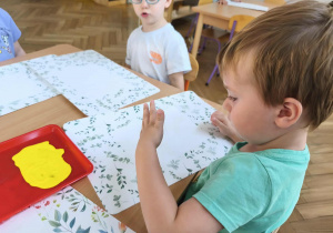 Na zdjęciu widzimy chlopca, który przygląda się swojej kolorowej dłoni odciśniętej w żółtej farbie.