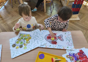 Na zdjęciu widzimy dwójkę dzieci, które z wykorzystaniem stempla w kształcie kropki wykonuje prace plastyczną.