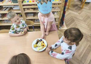 Na zdjęciu widzimy dziewczynkę, która częstuje dzieci owocami.