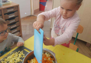 Zdjęcie przedstawia dziewczynkę zgarniającą pokrojone warzywa z deski do miski.