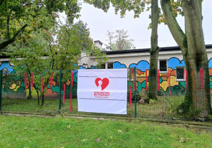 Zdjęcie przedstawia baner powieszony na ogrodzeniu przedszkola. Baner informuje o przystąpieniu przedszkola do kampanii Dzieciństwo bez przemocy.