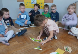 Dzieci siedzą w kole, a dziewczynka układa układankę na podłodze.