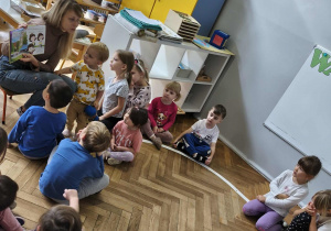 Na zdjęciu widzimy grupkę dzieci, które słuchają czytanej opowieści oraz oglądają ilustrację w książce.