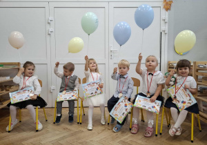 pasowane dzieci siedzą na krzesłach, trzymają dyplomy i balony
