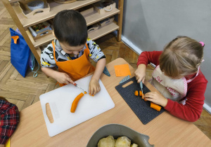 Na zdjęciu widzimy dwójkę dzieci, które kroją na deskach marchewkę do sałatki jarzynowej.
