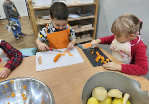 Na zdjęciu widzimy dwójkę dzieci, które kroją na deskach marchewkę do sałatki jarzynowej.