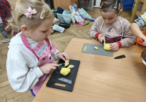 Na zdjęciu widzimy dwie dziewczynki, które kroją ziemniaki na deskach.