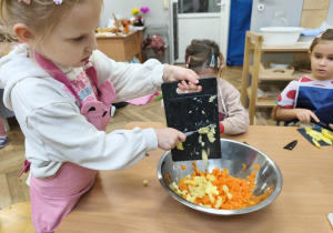 Na zdjęciu widzimy dziewczynkę, która zsuwa do miski z pokrojoną marchewką ziemniaka.