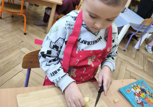 Na zdjęciu widzimy chłopca, który kroi kawałki jabłka na desce.