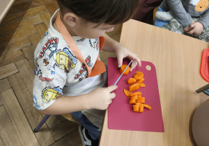 Na zdjęciu widzimy chłopca, który kroi na desce marchewkę do sałatki jarzynowej.