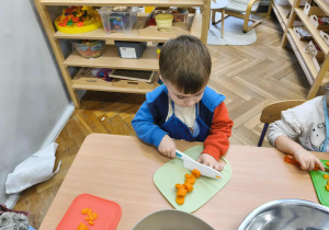 Na zdjęciu widzimy chłopca, który kroi na desce marchewkę do sałatki jarzynowej.