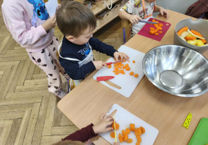 Na zdjęciu widzimy chłopca, który kroi na desce marchewkę do sałatki jarzynowej.