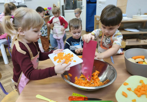 Na zdjęciu widzimy dziewczynkę i chłopca, którzy zsuwają z deski pokrojoną marchewkę do wspólnej miski.
