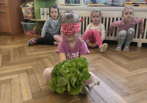 Zdjęcie przedstawia dziewczynkę, która próbuje rozpoznać sałatę po dotyku, z zasłoniętymi oczami.
