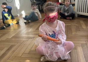 Zdjęcie przedstawia dziewczynkę, która próbuje rozpoznać marchewkę po dotyku, z zasłoniętymi oczami.