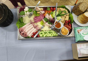 Na zdjęciu widzimy przygotowany dla dzieci stół szewdzki. Na półmisku znajdują się różne wędliny, sery, pomidorki, ogórek, kiełki rzodkiewki, sałata, kabanosy, kawałki pasztetu i inne produkty. Obok półmiska leży talerz z chlebami - białym i ciemnym razowym.